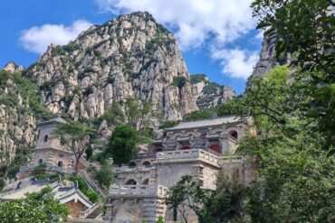 Chinese-style temple complex perched on a stone terrace against towering rocky cliffs, with red windows and carved stone railings amidst green trees.Sanhuangzhai,Shaolin & Songshan Area,Henan,Chinatravel