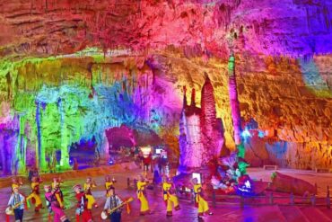 Colorfully lit cave interior with towering rock formations as a group of dancers in yellow traditional outfits perform with musicians in front of stalagmites.Jiuxiang Scenic Area,Kunming,Yunnan ,Chinatravel