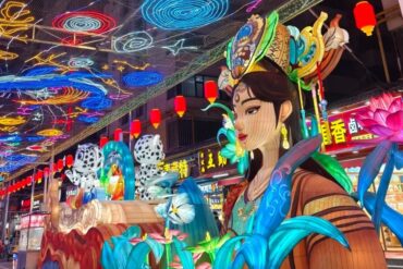 Vibrant Chinese lantern festival scene with a life-size woman statue wearing an ornate headpiece, surrounded by oversized blue leaves and pink lotus flowers beneath a ceiling of colorful neon lanterns.Mojia Subdistrict Night Market,Xining,Qinghai