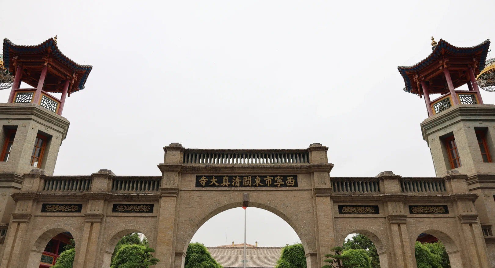 Stone arch gateway in a temple complex, with a central arch flanked by two towers featuring red, pagoda-style roofs and gold Chinese characters on plaques beneath.Dongguan Mosque,Xining,Qinghai
