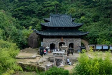 Traditional stone temple with dark tiled, multi-tiered roof nestled in a dense green forest; visitors at the entrance.Sanqing Mountain Scenic Area,Shangrao,Jiangxi,Chinatravel