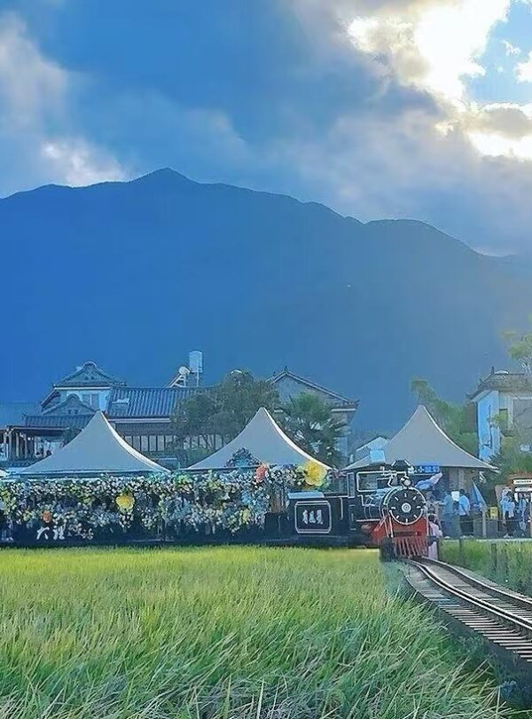 Steam locomotive at a countryside festival on a railway track, with white flower-decorated tents and a crowd, mountains in the distance.Xizhou and Zhoucheng Bai Villages,Dali,Yunnan,Chinatravel