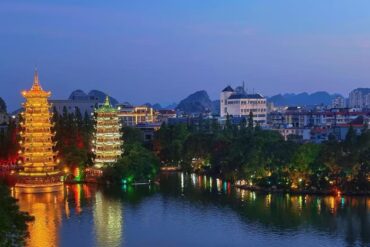 Two illuminated tiered pagodas beside a calm lake, reflections glowing gold and green with a cityscape in the background.Sun and Moon Twin Pagodas,Guilin,Guangxi ,Chinatravel