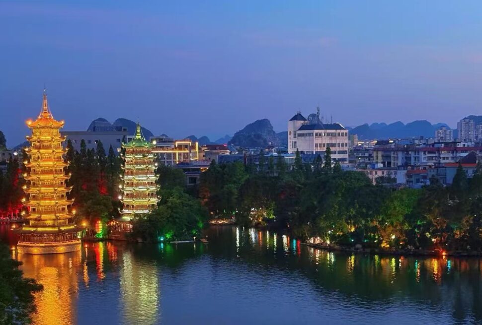 Two illuminated tiered pagodas beside a calm lake, reflections glowing gold and green with a cityscape in the background.Sun and Moon Twin Pagodas,Guilin,Guangxi ,Chinatravel