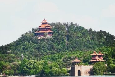 Red multi-tiered pagodas atop a forested hill beside the lake, with a stone gate at the shore in the foreground.Moshan Hill & Botanical Gardens,Wuhan,Hubei ,Chinatravel