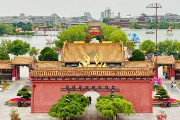 Dragon Pavilion Park & Park,Kaifeng,Henan,Chinatravel Red-walled Chinese temple entrance with golden tiled roofs and a central arch, leading to a tree-lined courtyard and pond in the distance.