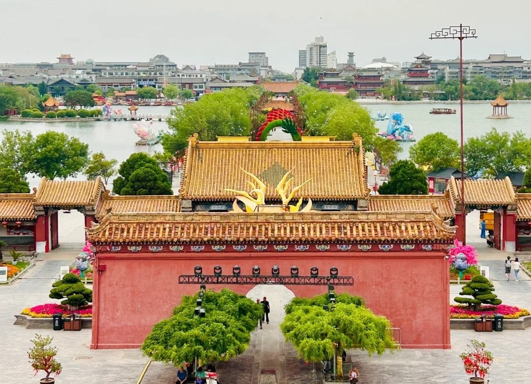 Dragon Pavilion Park & Park,Kaifeng,Henan,Chinatravel Red-walled Chinese temple entrance with golden tiled roofs and a central arch, leading to a tree-lined courtyard and pond in the distance.
