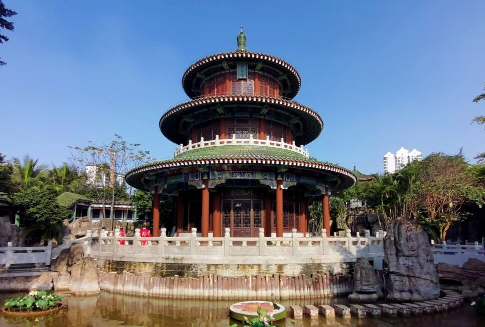 Hairui Tomb,Haikou,Hainan,Chinaexplorertour Three-tiered circular Chinese pavilion with red columns and green-tiled roofs in a garden, surrounded by a stone railing and a pond in the foreground.