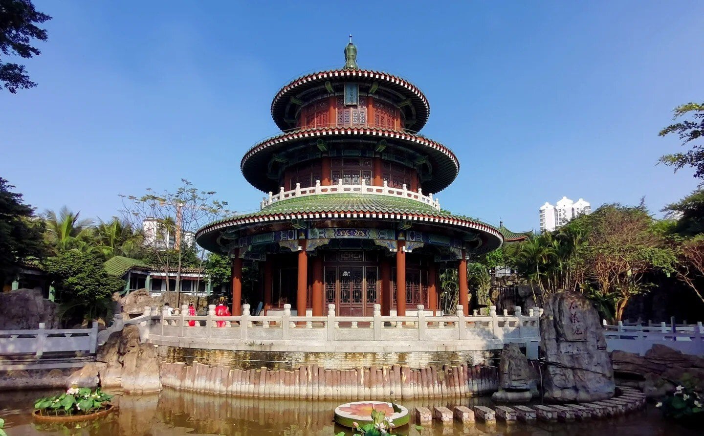 Hairui Tomb,Haikou,Hainan,Chinaexplorertour Three-tiered circular Chinese pavilion with red columns and green-tiled roofs in a garden, surrounded by a stone railing and a pond in the foreground.