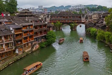 Fenghuang Ancient Town,Fenghuang,Hunan,Chinaexplorertour Ancient riverside town with wooden shophouses lining a green canal and a stone arch bridge, boats gliding on the water.