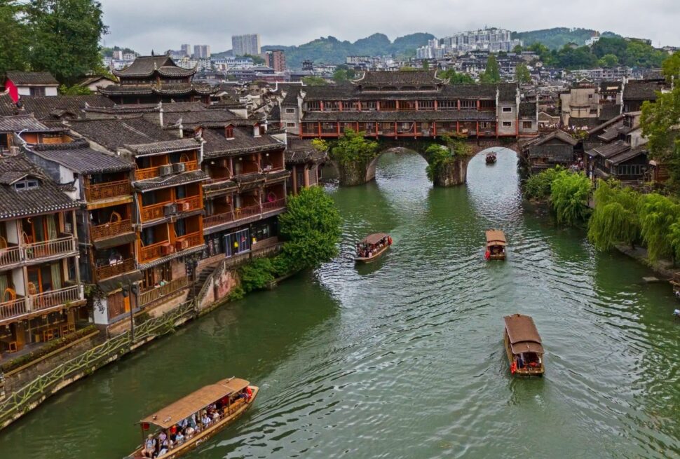 Fenghuang Ancient Town,Fenghuang,Hunan,Chinaexplorertour Ancient riverside town with wooden shophouses lining a green canal and a stone arch bridge, boats gliding on the water.