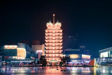 Erqi Memorial Tower,Zhengzhou,Henan,Chinaexplorertour Night view of a lit multi‑tiered pagoda tower in a city square, its warm glow reflected on the wet pavement.