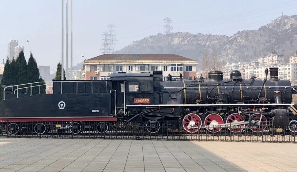 Dalian Modern Museum,Dalian,Liaoning,Chinatravel Historic steam locomotive on display in a public square; black body with red wheels and white piping, mountainous backdrop.