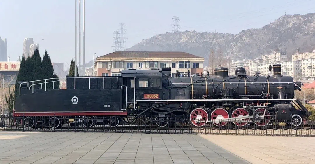 Dalian Modern Museum,Dalian,Liaoning,Chinatravel Historic steam locomotive on display in a public square; black body with red wheels and white piping, mountainous backdrop.