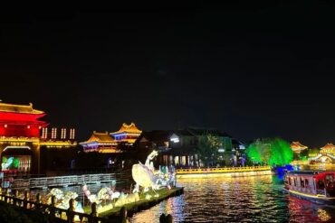Millennium City Park,Kaifeng,Henan,Chinatravel Night view of a lit riverside with traditional East Asian pagodas, red and gold buildings, and colorful lamp-lit structures reflecting on the water; a boat glides along the river.