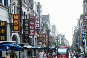 Beihai Old Street,Beiahi,Guangxi,Chinaexplorertour Busy street in China lined with tall vertical signs in Chinese characters and market stalls on both sides, with pedestrians and a red bus in the distance.