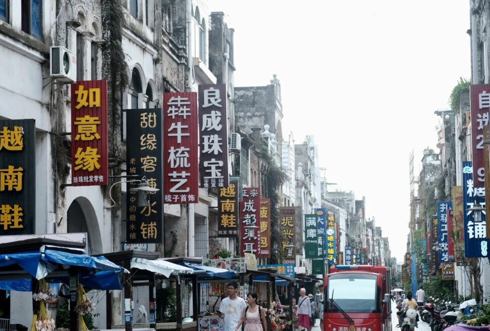 Beihai Old Street,Beiahi,Guangxi,Chinaexplorertour Busy street in China lined with tall vertical signs in Chinese characters and market stalls on both sides, with pedestrians and a red bus in the distance.