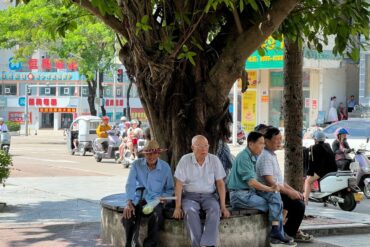 Jiaji Old Town,Qionghai,Hainan,Chinaexplorertour Elderly men sitting on a circular bench under a large tree in a busy urban street scene with scooters and storefronts in the background.