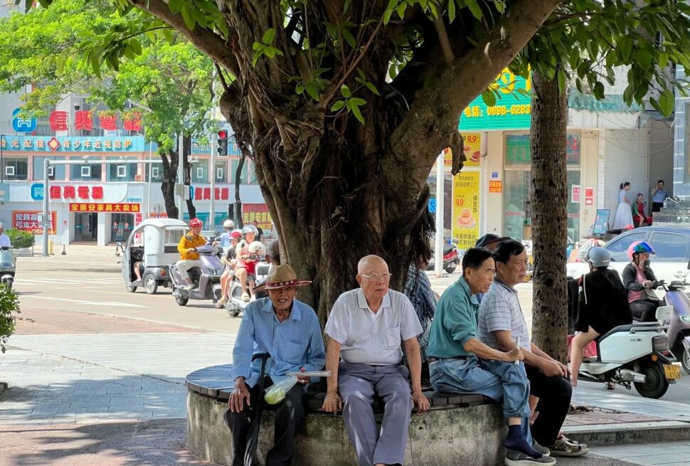Jiaji Old Town,Qionghai,Hainan,Chinaexplorertour Elderly men sitting on a circular bench under a large tree in a busy urban street scene with scooters and storefronts in the background.