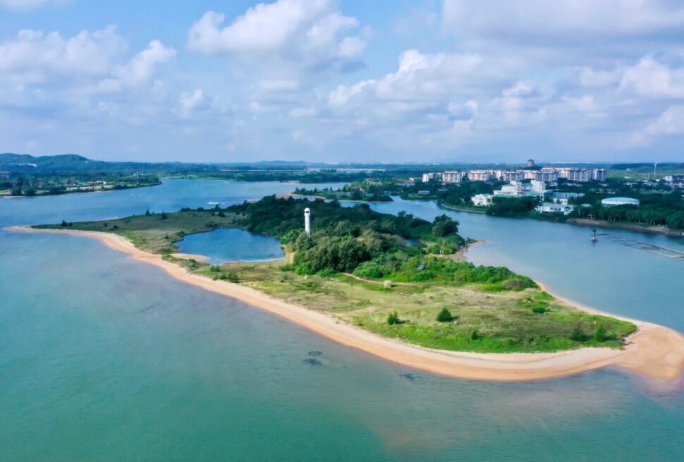Wanquan River Scenic Area,Qionghai,Hainan,Chinaexplorertour Aerial view of a small green island with a white lighthouse, sandy beaches, and surrounding blue water; a town is visible in the distance.
