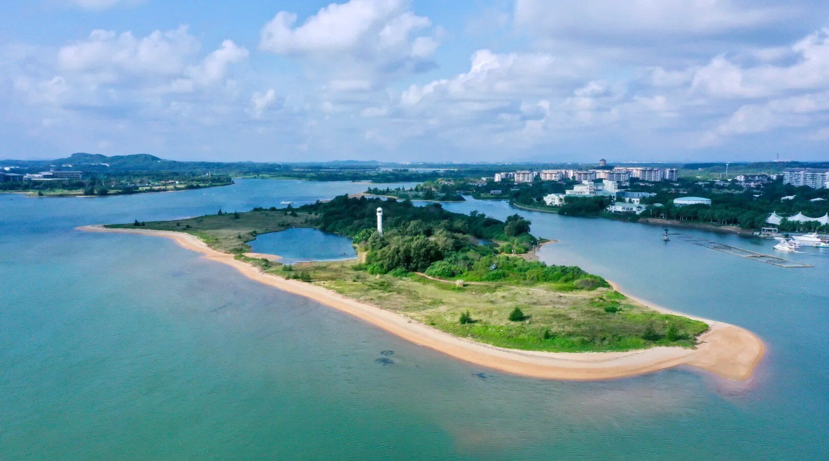 Wanquan River Scenic Area,Qionghai,Hainan,Chinaexplorertour Aerial view of a small green island with a white lighthouse, sandy beaches, and surrounding blue water; a town is visible in the distance.