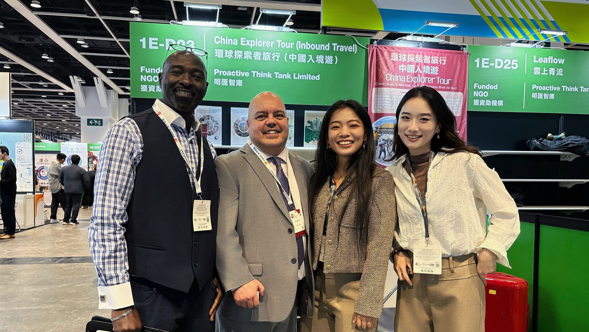 Four professionals pose for a group photo at a trade show booth with green signage overhead, smiling and wearing lanyards.
