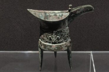 Zhengzhou Museum,Zhengzhou,Henan,Chinaexplorertour Ancient bronze ritual vessel with a long curved spout, standing on three legs, featuring ornate scrolling decorations around the middle.