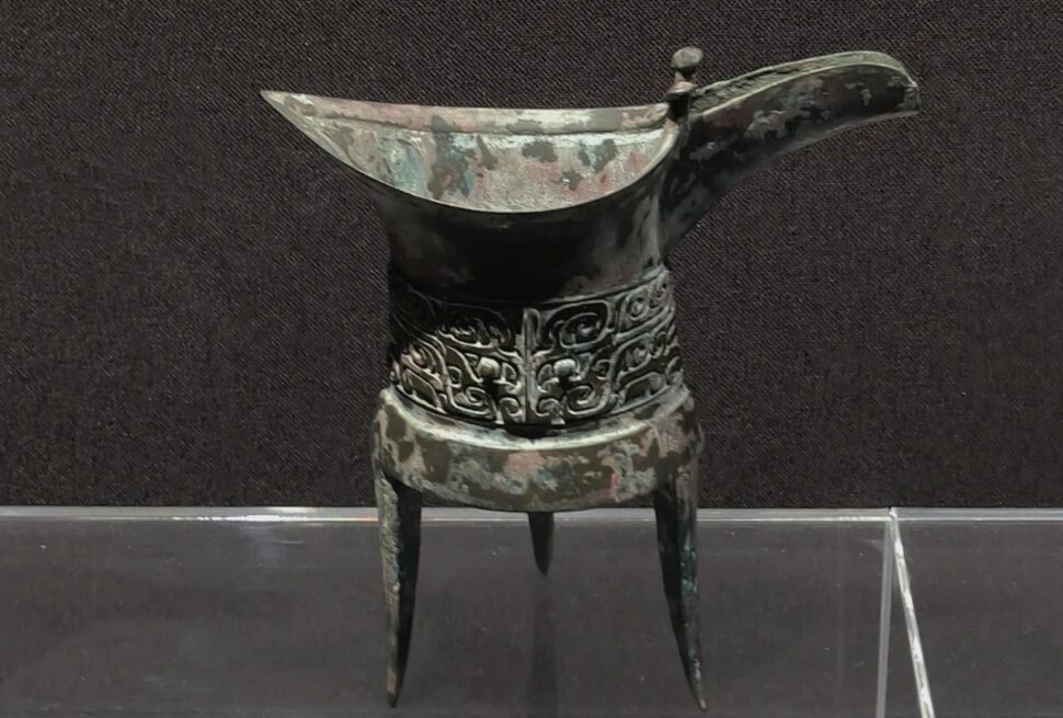 Zhengzhou Museum,Zhengzhou,Henan,Chinaexplorertour Ancient bronze ritual vessel with a long curved spout, standing on three legs, featuring ornate scrolling decorations around the middle.
