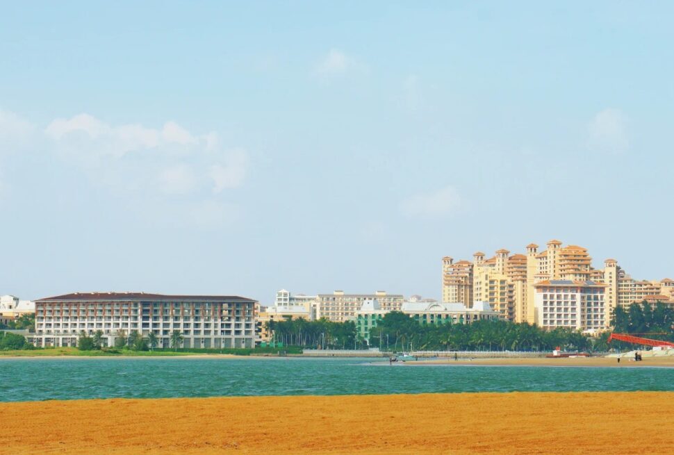 Bo’ao Jade Belt Beach,Qionghai,Hainan,Chinaexplorertour Beach scene with orange sand in the foreground, blue water, and cluster of resort hotels along the shore under a clear sky.