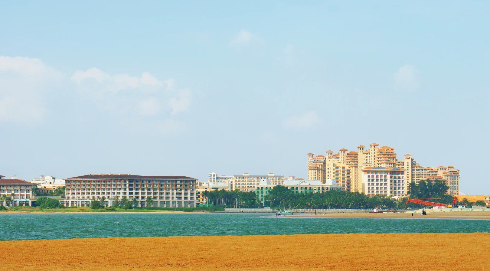 Bo’ao Jade Belt Beach,Qionghai,Hainan,Chinaexplorertour Beach scene with orange sand in the foreground, blue water, and cluster of resort hotels along the shore under a clear sky.