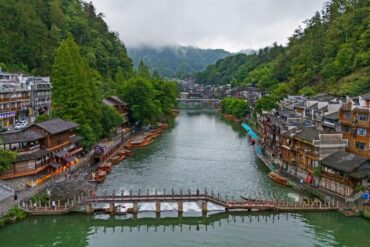 Qinglong Ancient Bridge & Miao Architecture,Fenghuang,Hunan,Chinaexplorertour River town along a calm river with boats docking at the banks and a stone bridge in the foreground. Lush green hills surround traditional multi-story buildings on both sides.