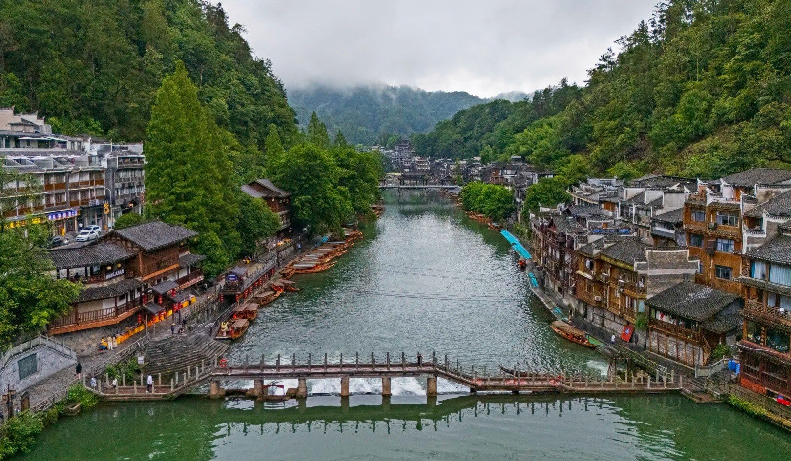 Qinglong Ancient Bridge & Miao Architecture,Fenghuang,Hunan,Chinaexplorertour River town along a calm river with boats docking at the banks and a stone bridge in the foreground. Lush green hills surround traditional multi-story buildings on both sides.