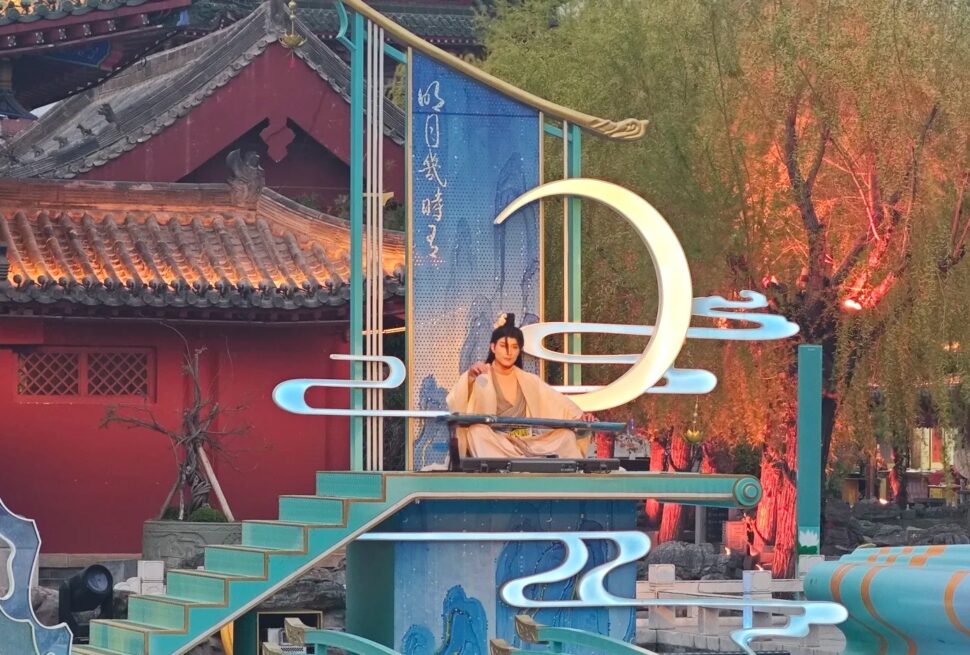 Kaifeng Mansion,Kaifeng,Henan,Chinaexplorertour Performer in traditional clothing sits on a turquoise stage with decorative neon shapes and a large illuminated crescent moon sculpture in a temple courtyard setting.