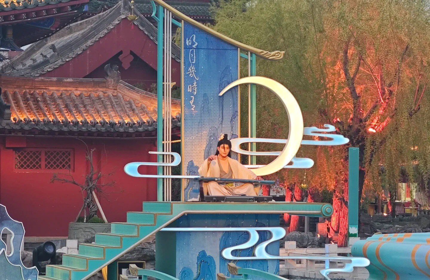 Kaifeng Mansion,Kaifeng,Henan,Chinaexplorertour Performer in traditional clothing sits on a turquoise stage with decorative neon shapes and a large illuminated crescent moon sculpture in a temple courtyard setting.