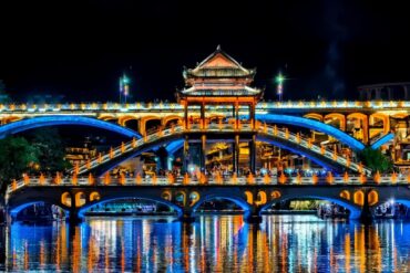 Fenghuang Ancient Town,Fenghuang,Hunan,Chinaexplorertour Night photograph of a pagoda-style bridge lit in blue and orange, spanning a calm river with colorful reflections and a central pavilion.