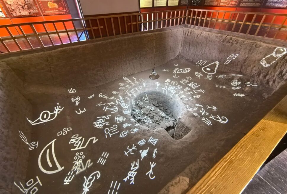 Yin Ruins (Yinxu, UNESCO),Anyang,Chinaexplorertour Indoor museum exhibit: a sunken rectangular pit with a central rock-filled hole, surrounded by white glyphs on the floor and a railing along the edge.