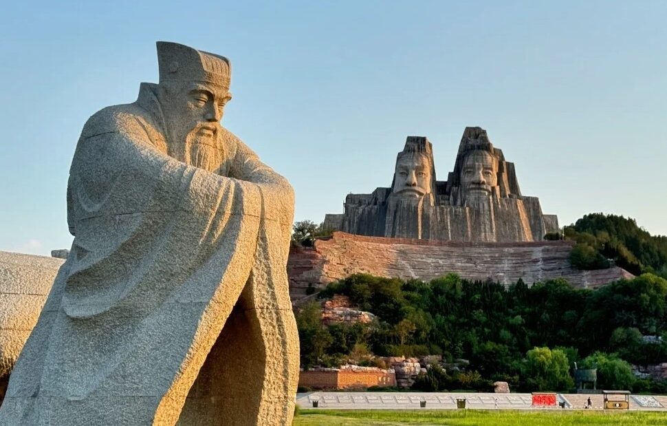 Yellow River Scenic Area,Zhengzhou,Henan,Chinaexplorertour Yellow River Scenic Area,Zhengzhou,Henan,ChinaexplorertourA large seated stone statue in a park with a multi-face rock sculpture carved into a hill in the background, surrounded by trees and landscaping.