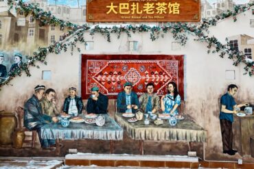 Grand Bazaar,Urumqi,Xinjiang,Chinaexplorertour Mural of a traditional tea-house scene: people dining at two long tables, with a red rug backdrop, greenery, and a sign reading 'Grand Bazaar old Tea House' (Chinese text above).