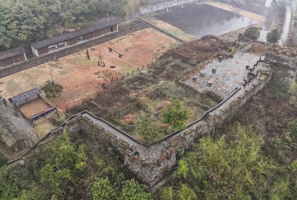 Southern Great Wall Ruins,Fenghuang,Hunan,Chinaexplorertour Aerial view of an old stone fortress with walls, stairways, and a central courtyard amid surrounding trees and hills.