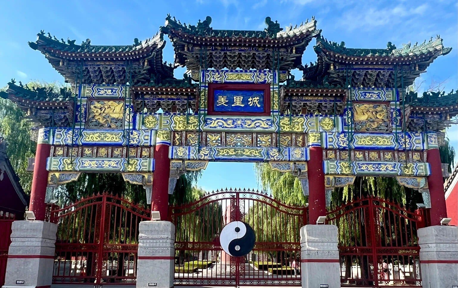Youli Castle & Scenic Area,Anyang,Henan,Chinaexplorertour Ornate Chinese archway with blue plaque reading '美里城', intricate gold carvings, red pillars, and a yin-yang symbol hanging in front of a gated courtyard.