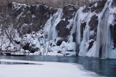Changbai Waterfall,Baishan,Jilin,Chinatravel Winter scene of a snow-covered cliff with several frozen waterfalls cascading into a blue lake, a bare-branched forest on the left, and a wooden boardwalk along the shore.