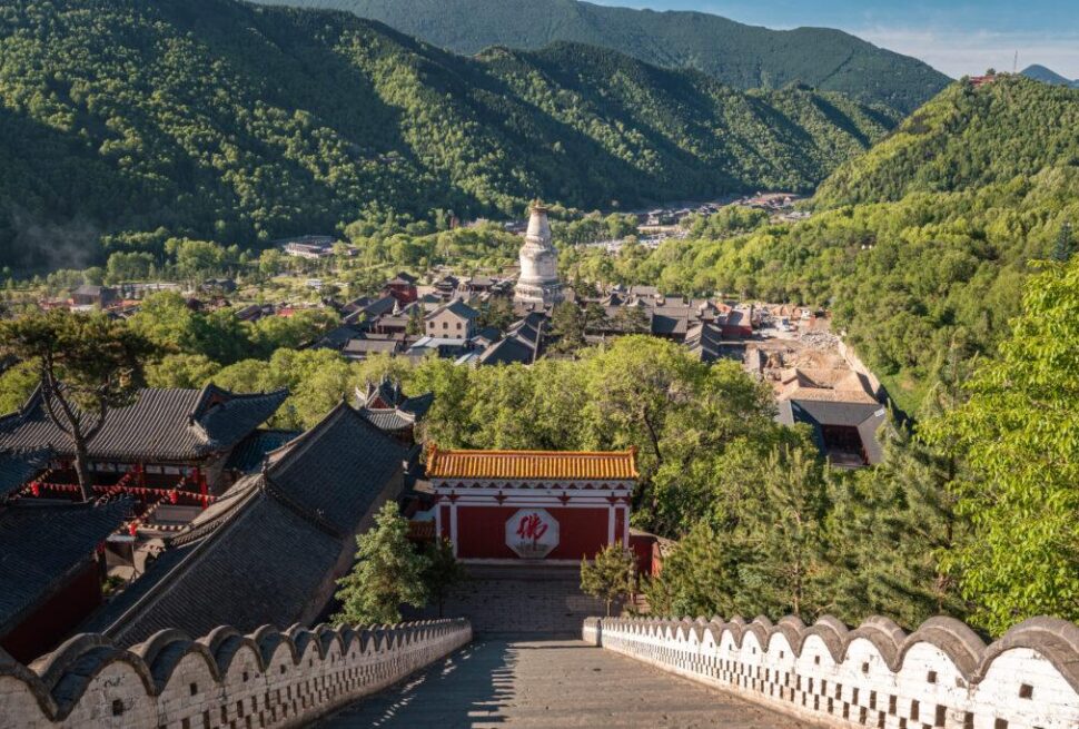 Wide view of a temple complex nestled in green mountains, with a long stone staircase descending toward red gatehouses and traditional roofs.