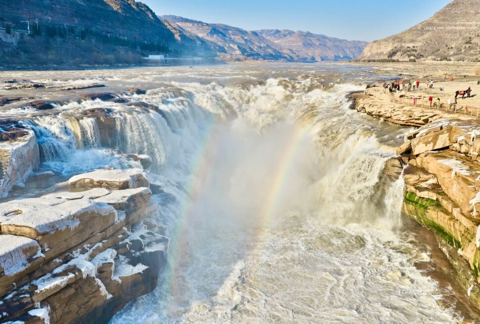 Wide waterfall with mist and a small rainbow, rocky cliffs, and people on a viewing platform to the right,Hukou Waterfall,Linfen,Shanxi