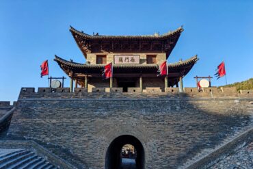 Ancient Chinese city gate with a curved tiled roof and brick walls, red banners on the battlements, and a stone archway beneath a clear blue sky,Yanmen Pass,Xinzhou,Shanxi