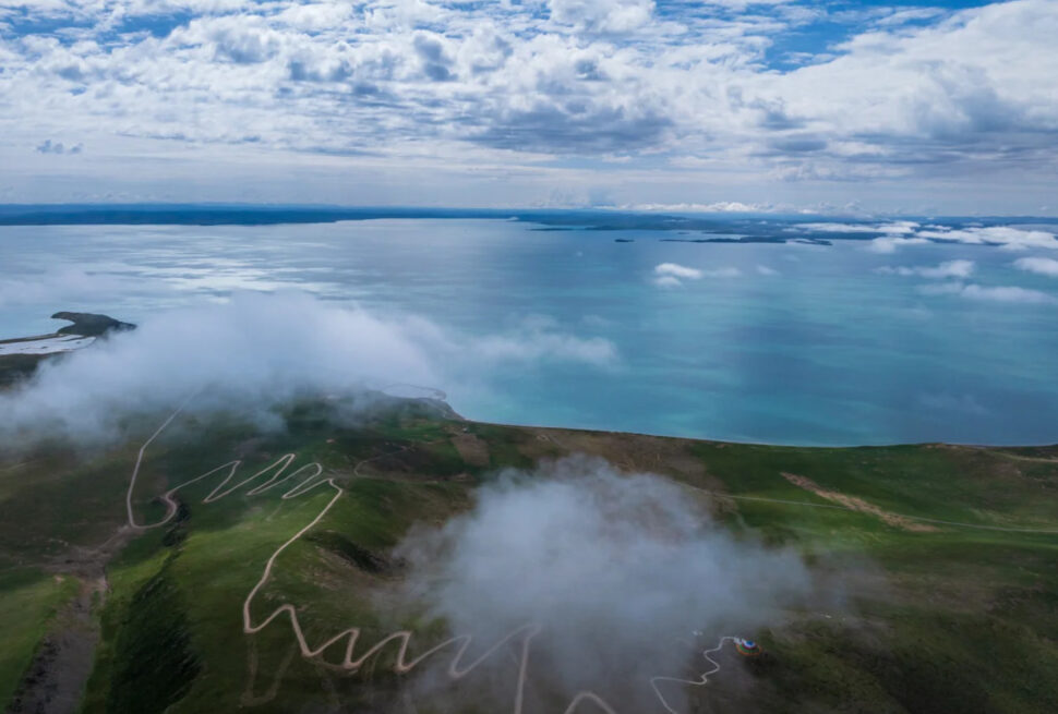 Aerial view of turquoise sea along green hills, with a winding road and cloudy sky above the coastline. Better alt text would be concise and informative for screen readers.Sanjiangyuan National Park,Yushu, Qinghai