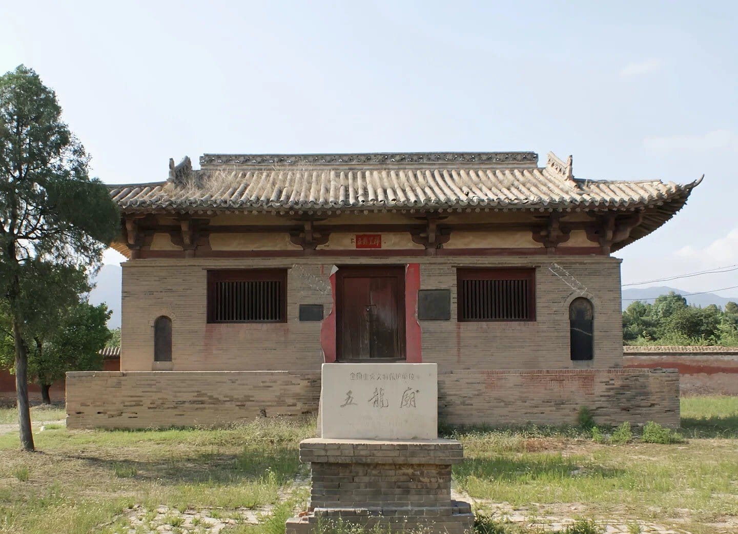 Traditional Chinese brick building with a tiled, curved roof and red central doorway, set in a grassy yard with a stone pedestal in front.Guangrenwang Temple,Yuncheng,Shanxi