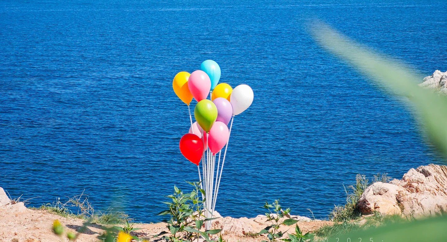 A colorful bunch of balloons on a rocky coastline overlooking the blue sea.