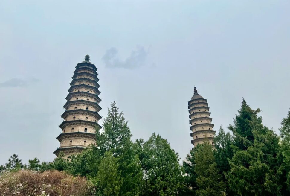 Two tiered stone pagodas rise among green trees against a pale blue sky, creating a tranquil scene.”Shuangta Temple,Taiyuan, Shanxi