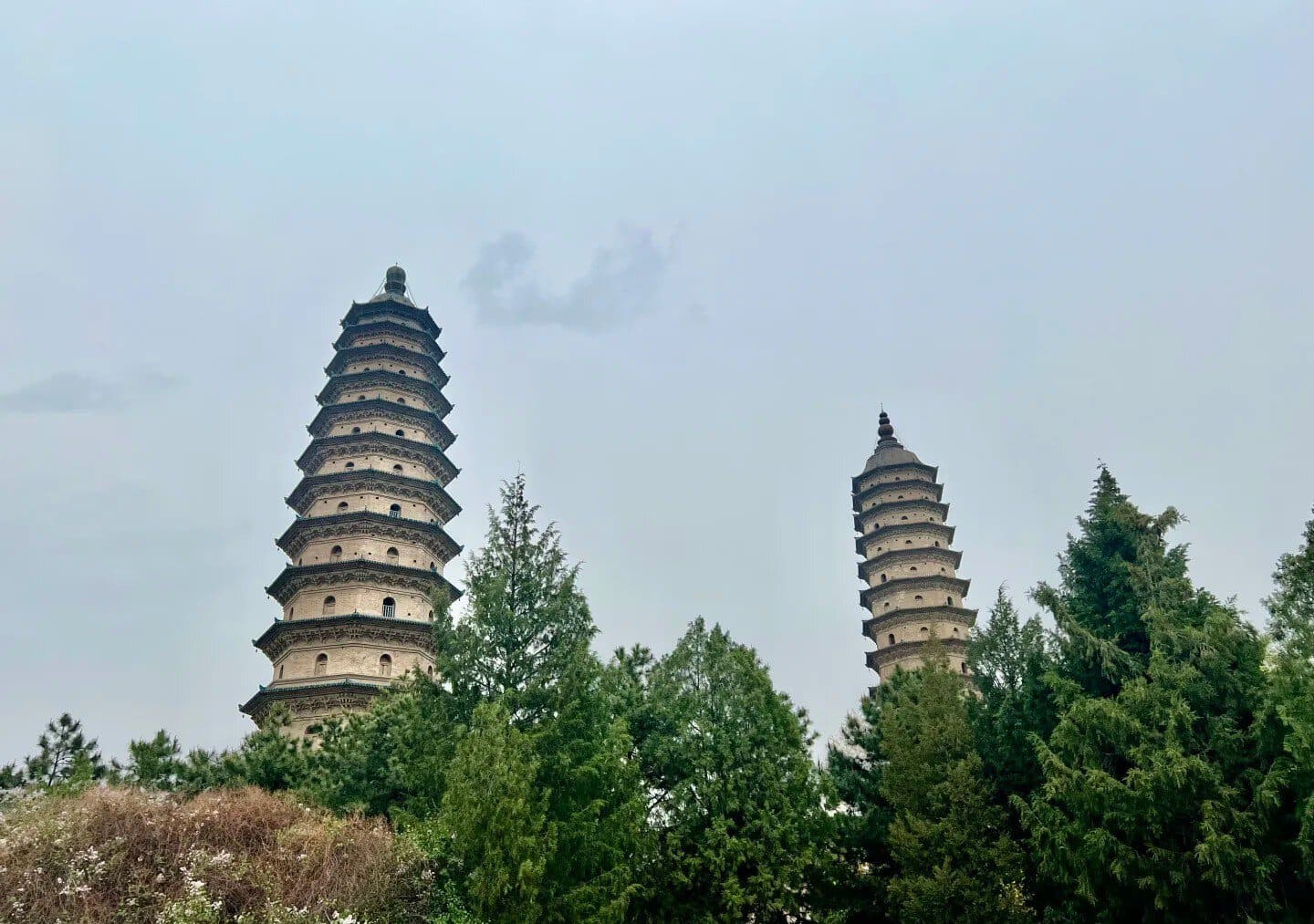 Two tiered stone pagodas rise among green trees against a pale blue sky, creating a tranquil scene.”Shuangta Temple,Taiyuan, Shanxi