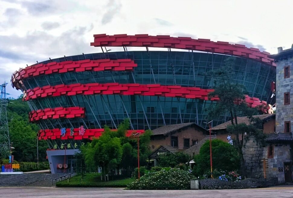 Futuristic circular glass building wrapped in red horizontal panels beside a stone tower and greenery.Yeyuhai Tourist Resort,Liupanshui,Guizhou